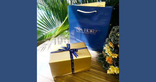 Blue gift bag with 'NA HOKU' branding next to a gold gift box with a blue ribbon on a wooden surface.
