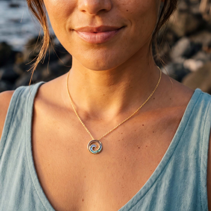 A close-up lifestyle shot of a woman wearing a gold ocean wave pendant featuring abalone and Koa wood inlay on a gold chain, set against a blurred outdoor rocky background. Item No. 45273