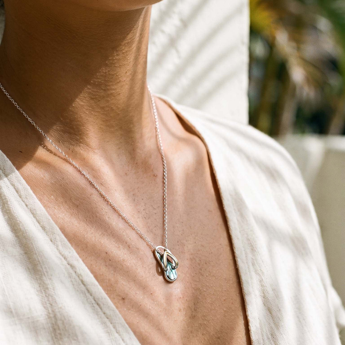 A close-up lifestyle shot of a person wearing a delicate silver chain with a silver and gold slipper pendant featuring abalone and Koa wood inlay. Item No. 45404