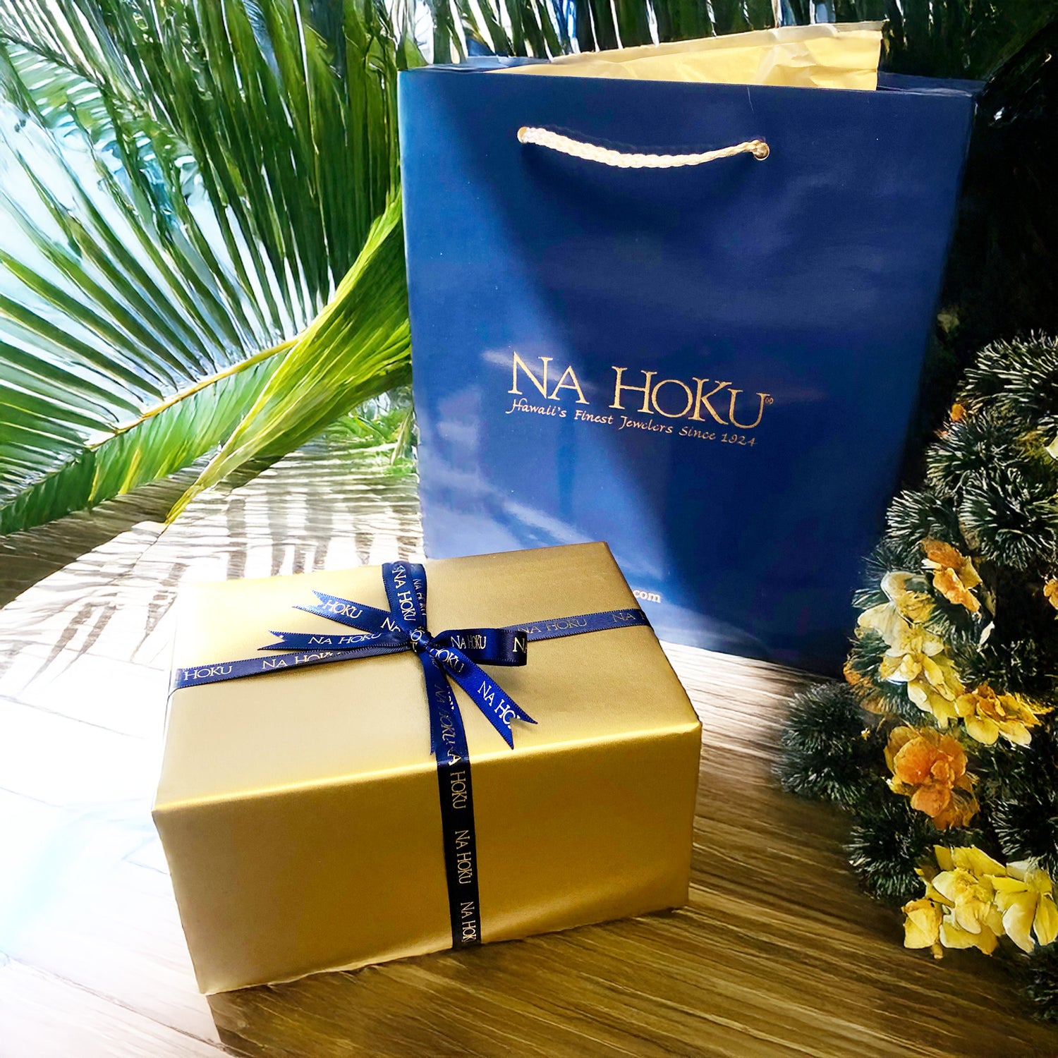 Na Hoku gift bag and gift wrapped box with ribbons sitting on wooden table with palm leaves and small Christmas tree with flowers